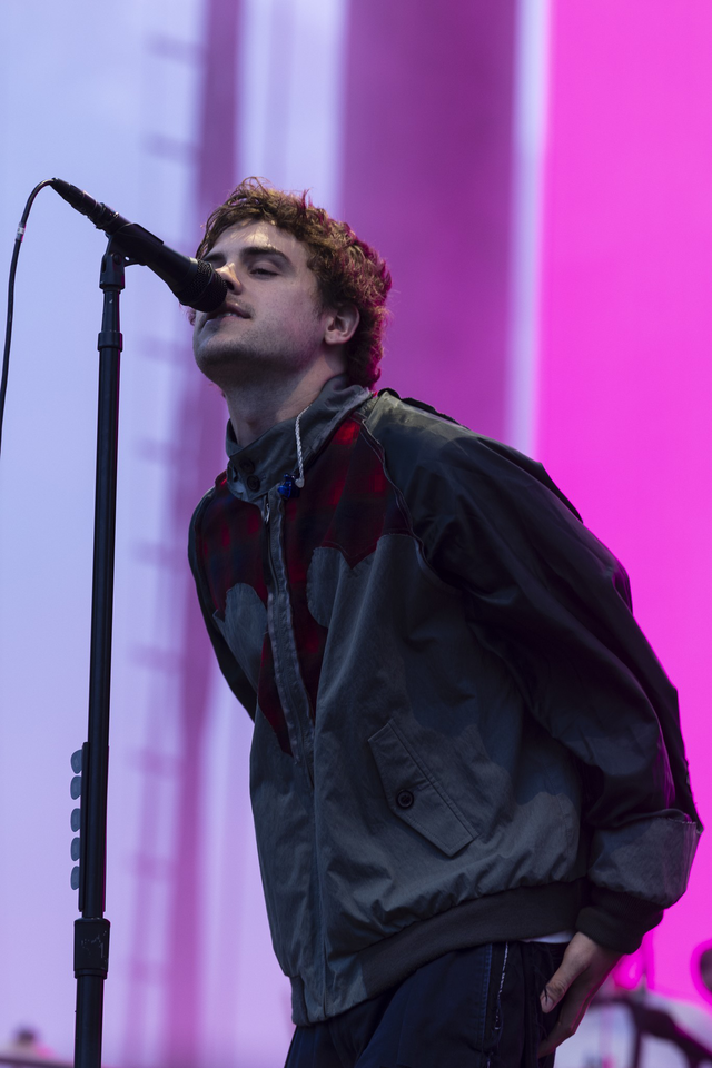 A man with curly hair is singing into a microphone on stage, wearing a gray and red jacket against a bright pink and purple background.